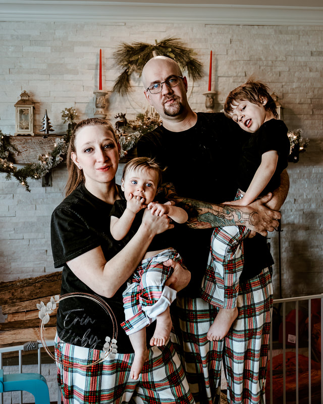 An image of a family of four. A mother holds an infant on the left side of the frame. Towards the center and back a father stands, holding his son who is on the right side of the image. The family wears black tee shirts and white/green/red plaid pj bottoms. They stand in front of a fireplace with a large, white stone facade.