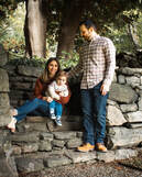 A mother in a rust colored sweater and blue jeans holds a toddler in a cream sweater and blue jeans, while sitting on stone steps. A father stands in front of them and to the left, looking down at the woman and toddler, with his hand on her shoulder. The stone steps lead up to trees and a garden. A stone path lies at the bottom of the steps.