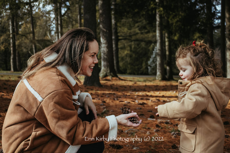 Background depicts a pine forest. Pine needs and pine cones blanket the ground. In the foreground a mother wearing a brown coat with white trim hands a pinecone for her daughter on the left. The mother has long brown hair. The daughter is a toddler, has light brown hair tied up with a red ribbon, and has a tan coat. 